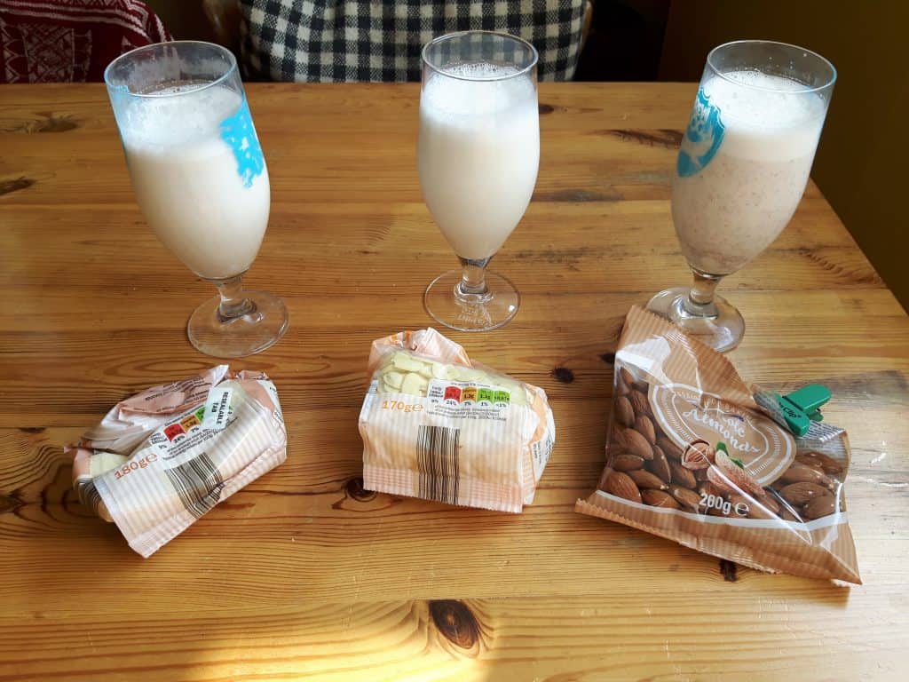 Three glasses of almond milk made with three different types of almond; ground, flaked and whole; on a wooden table.